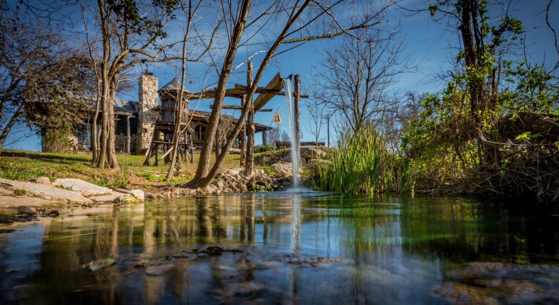 A serene pond with a small waterfall in front of a stone building surrounded by trees.