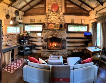 Interior view of a log cabin's brick fireplace and sitting area with kitchen in background.