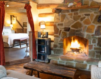 View across living room with gray furniture toward bedroom, fireplace burning in stone enclosure, flat television on wall in corner.