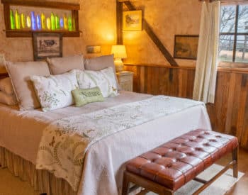 Cream painted and wood paneled bedrooms with queen bed with white quilt and lots of pillows, wooden shelves, stand with lit lamp in corner, window with curtains