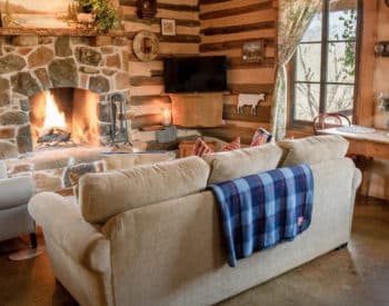 Living room with cream cushioned furniture, fireplace burning in stone setting, wooden walls, pine table with 2 chairs by windows with curtains, flat tv on stand in corner.