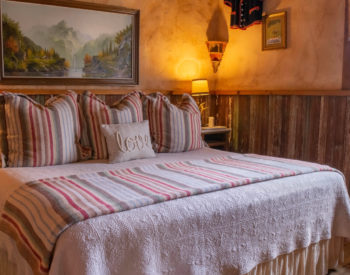 Bedroom with painted walls with wooden paneling, artwork on walls, stands with lamps by bed with white comforter and striped pillows, wooden floor