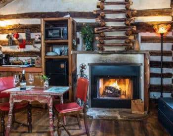 Kitchenette with refrigerator, microwave and dishes. Small table with red cushioned chairs, burning fireplace, lamp in corner, blue wooden bench under window.