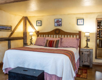 Cream painted bedroom with full bed with oak headboard, black bench at end of bed, pictures on walls, nightstands with lamps