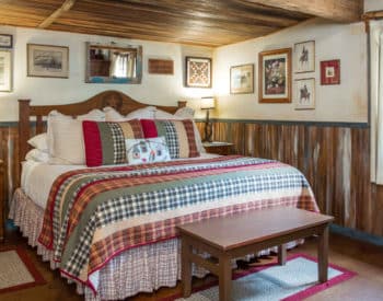 High bed with checkered comforter and bed skirt, pictures on walls, window in shape of heat, wood bench.