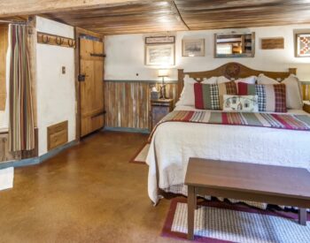 A country style bedroom with wood wainscotting, a bed with a table at the foot of the bed, and a corner tub and shower.