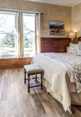 A country style bedroom with wood floors, wood wainscotting, a bed, white wood nightstands with lamps, a green armchair and ottoman, and a fireplace in the fire next to a large window.