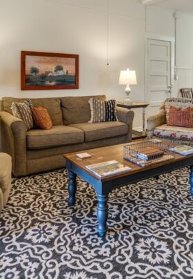 A living room and dining area with wood floors, a large area rug, a sofa and plush armchair, a wood coffeetable.