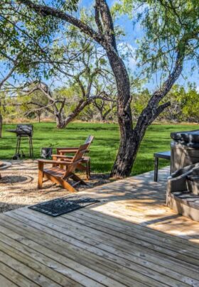 An inviting outdoor area with a wood deck and hot tub, chairs and a bench surrounding a fireplace, green grass, trees, and blue skies.