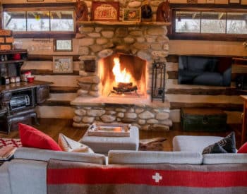 Gray couch with blanket draped over back, Burning fireplace in stone enclosure, kitchenette on left, table for two by window on right