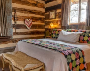 Queen bed with white coverings & colorful blanket with two windows with curtains, nightstands on sides with lamps, cushioned bench at foot,