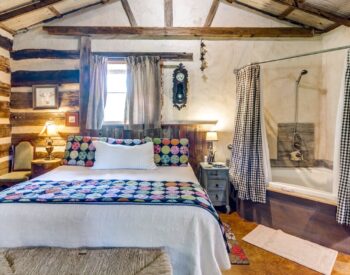 A country bedroom with log walls on 2 of the walls, wood beams on the ceiling, a bed, and a corner tub.