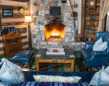 Living rooms with blue leather furniture with throw pillows and blue blanket, burning fireplace in stone enclosure, table for 2 on left bedroom through curtains on right.