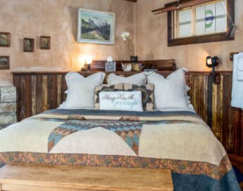 Bedroom with queen bed with cream walls with wood at bottom, towels hanging on racks, elevated bathtub on right in wooden frame.