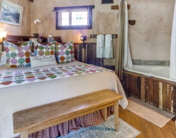 A country style bedroom with a bed, wood accents, an antique metal fireplace, and a corner tub.