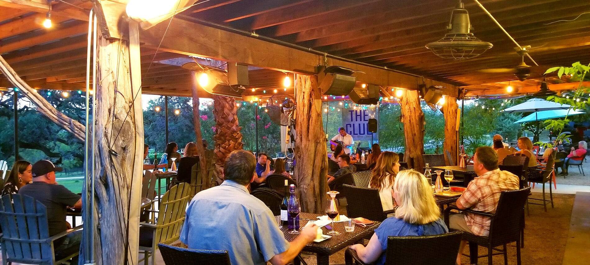 Group of visitors sitting at brown square tables under covered patio with hanging lights listening to live entertainment.