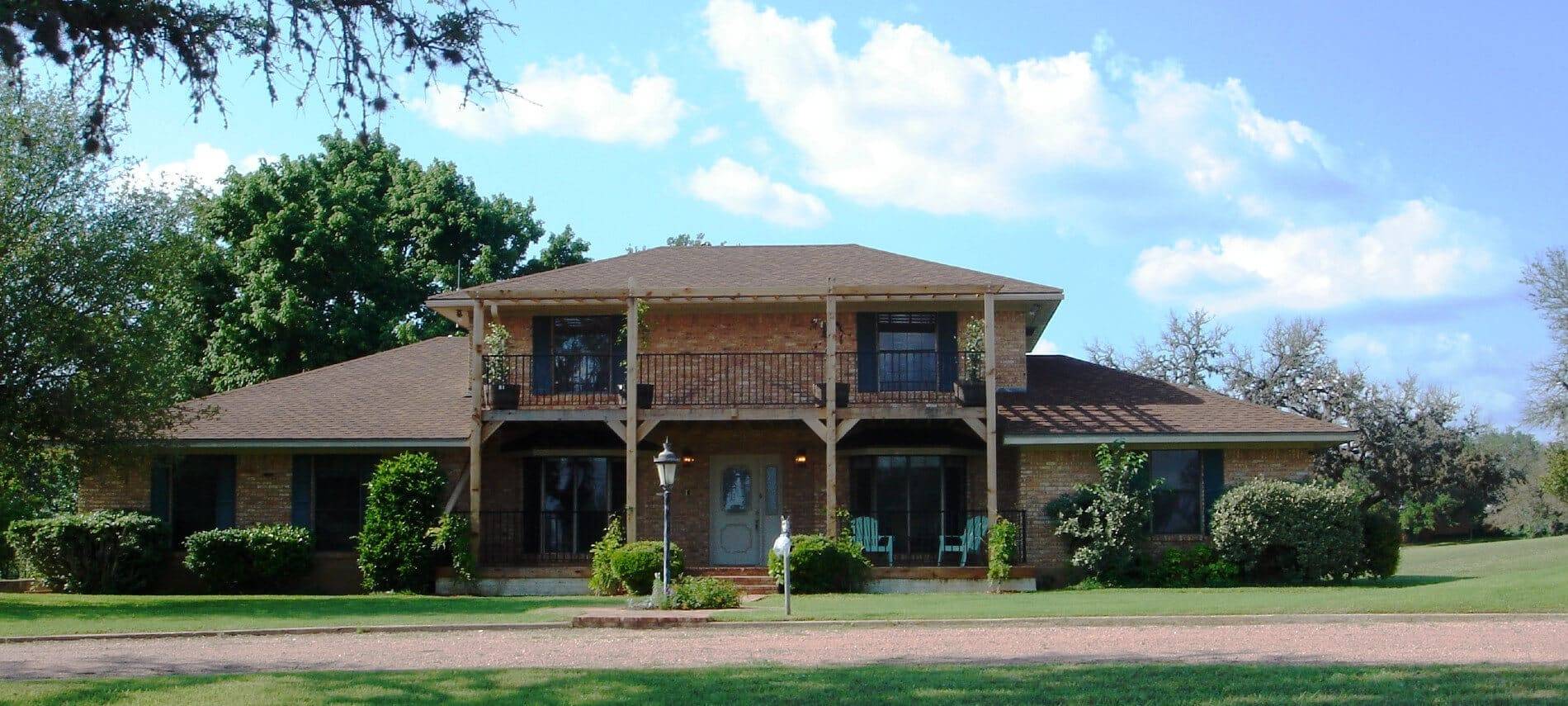 two-story tan brick house with 2nd floor balcony, bushes in front surrounded by trees, porch with green chairs.