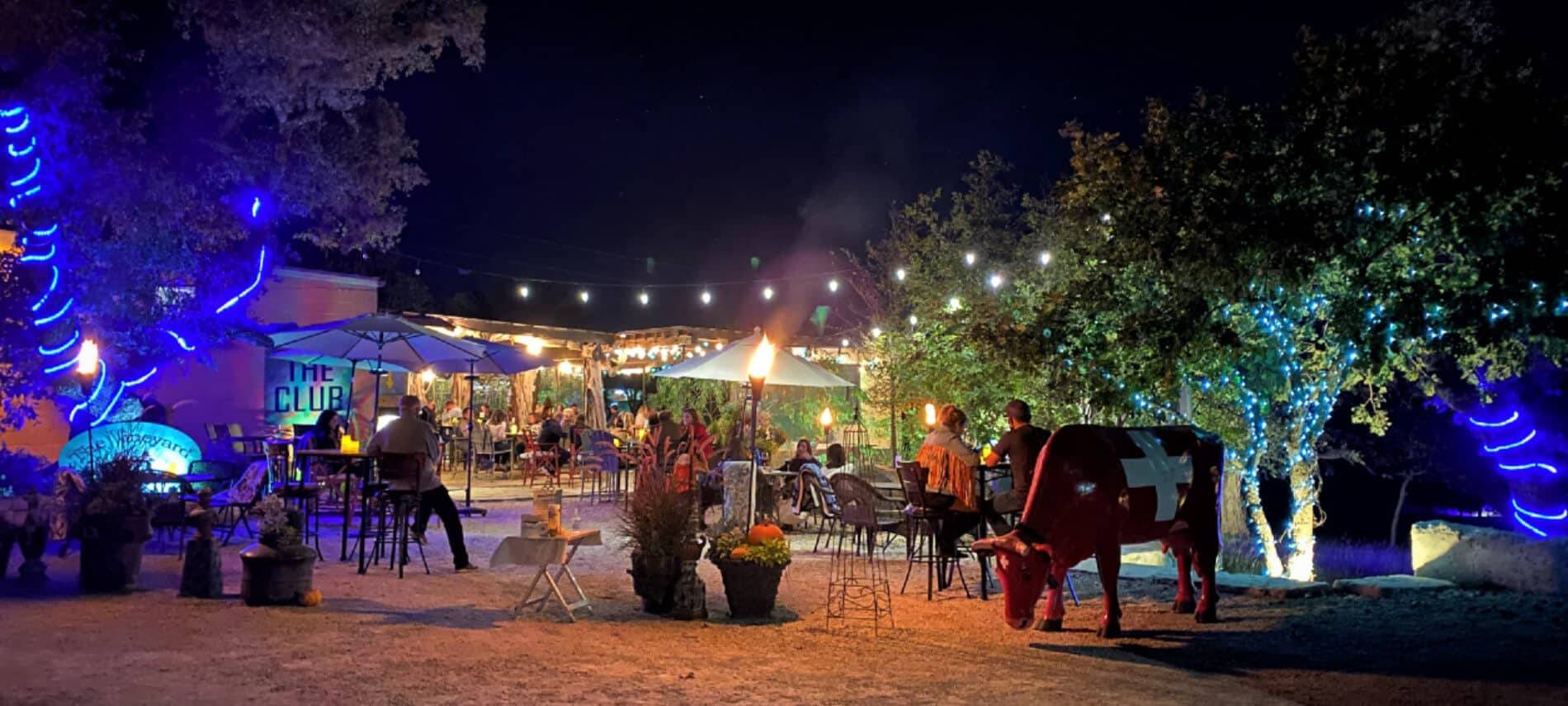 Patio Seating at The Club at night with hanging lights and tables with umbrellas. Red cow figurine with white cross.
