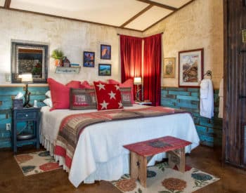 Queen bed with white covering and numerous pillows, wood bench at foot, wooden brown and cream walls, elevated bathtub with wooden frame.