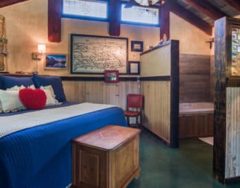 Blue covered bed with numerous pillows, oak bed at foot, view toward wooden framed tub on right.